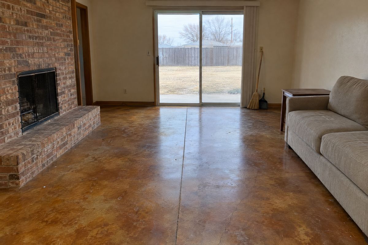 Den with a mottled amber and brown acid-stained concrete floor, a visible control joint and hairline crack, a broom by a sliding door and a sofa.