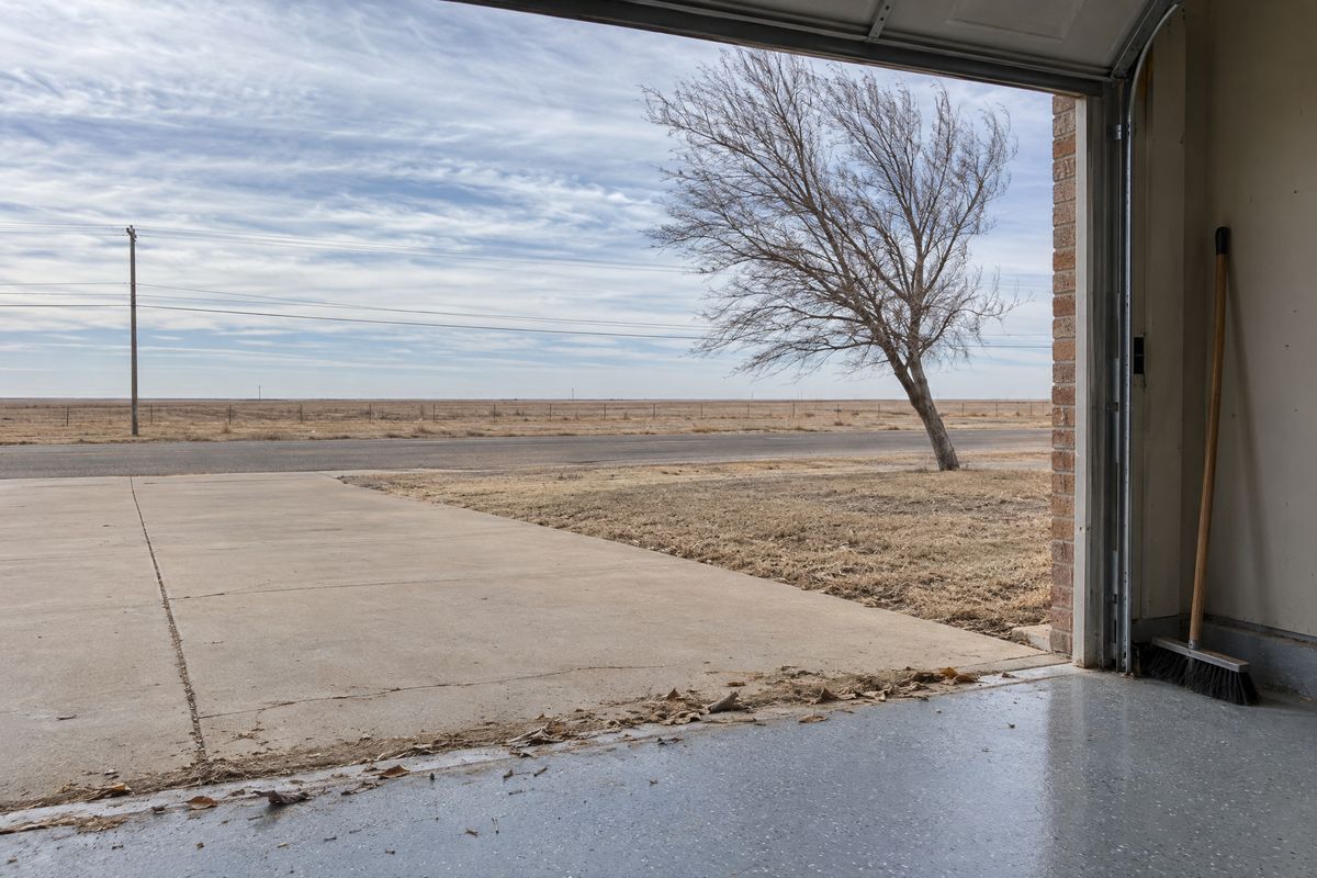 View out of an open garage on a windy winter day, blown dust along the threshold of a gray flake floor, a broom and a bare elm bending outside.