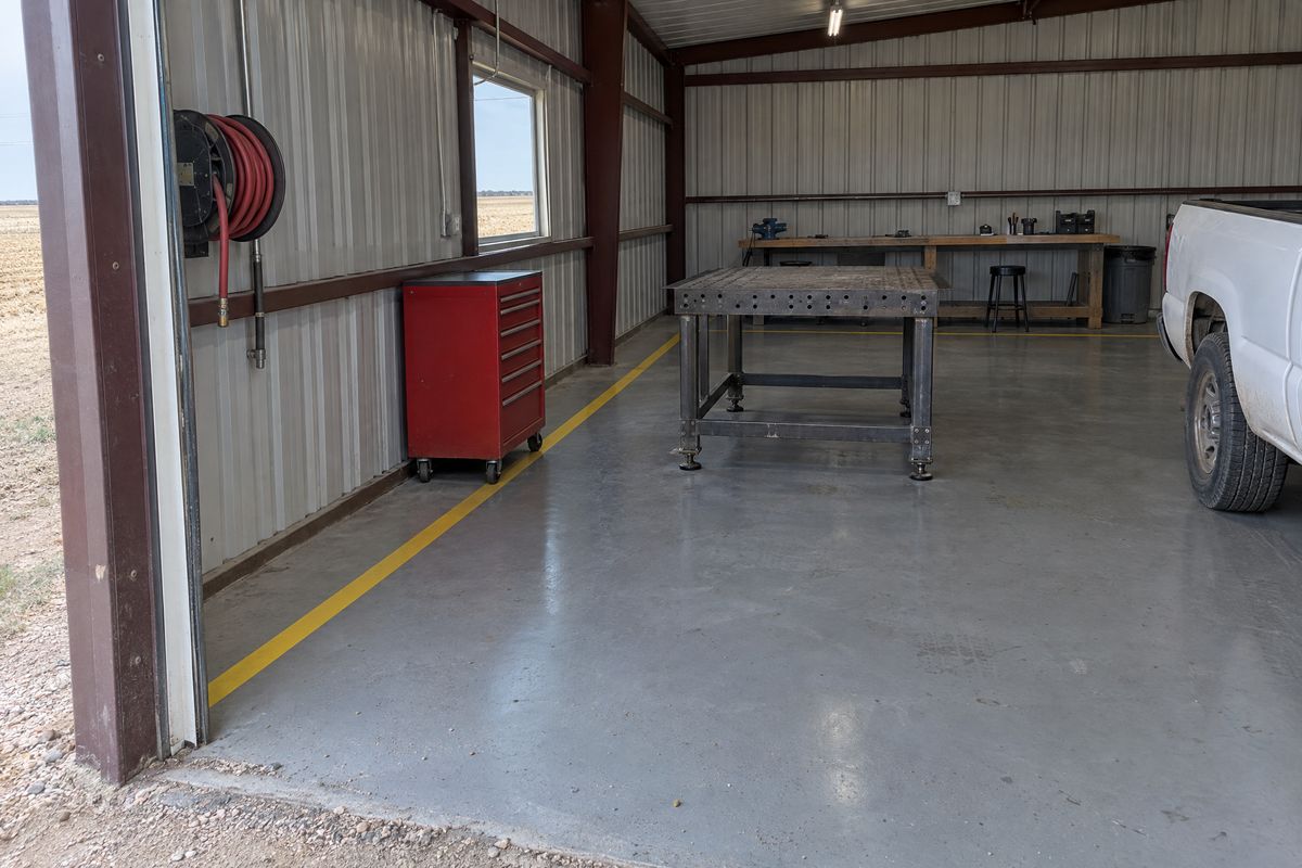 Metal farm shop with a gray epoxy floor and yellow walkway lines, a welding table, a red tool chest, a workbench and a pickup's side at the edge.