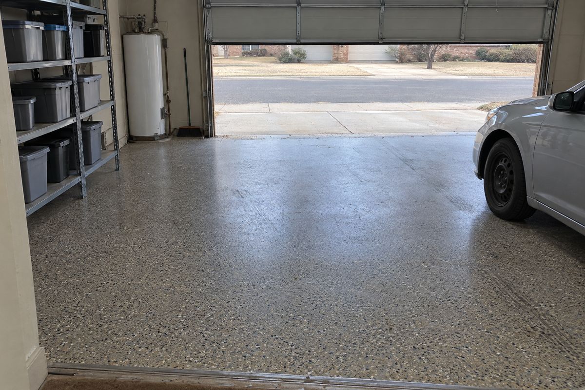 Attached two-car garage with a gray, black and tan flake floor, faint tire marks, metal shelving with bins, a water heater and a sedan's front fender.