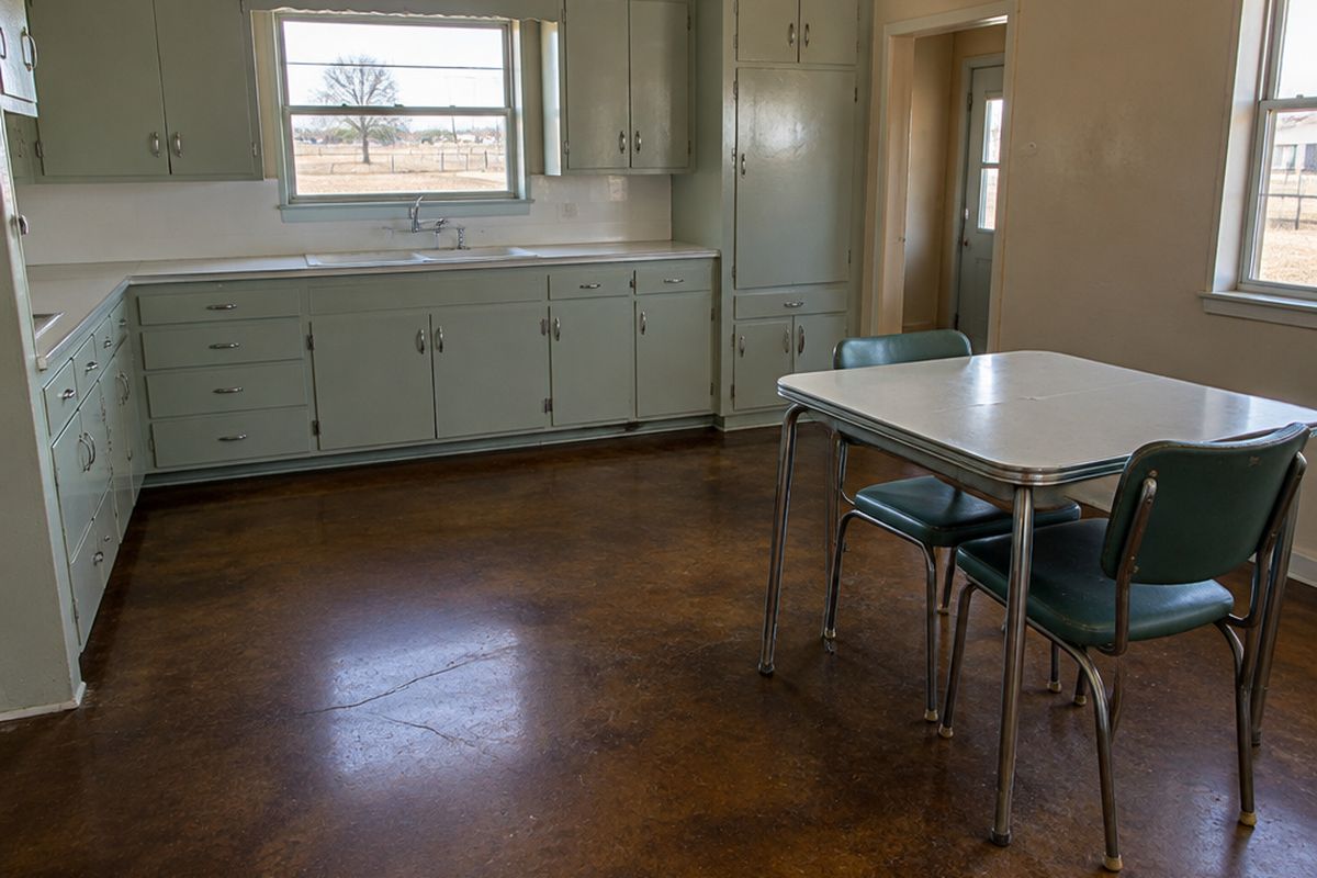 Kitchen and dining area with a dark brown and amber stained concrete floor showing faint craze cracks, painted cabinets and a small table with chairs.
