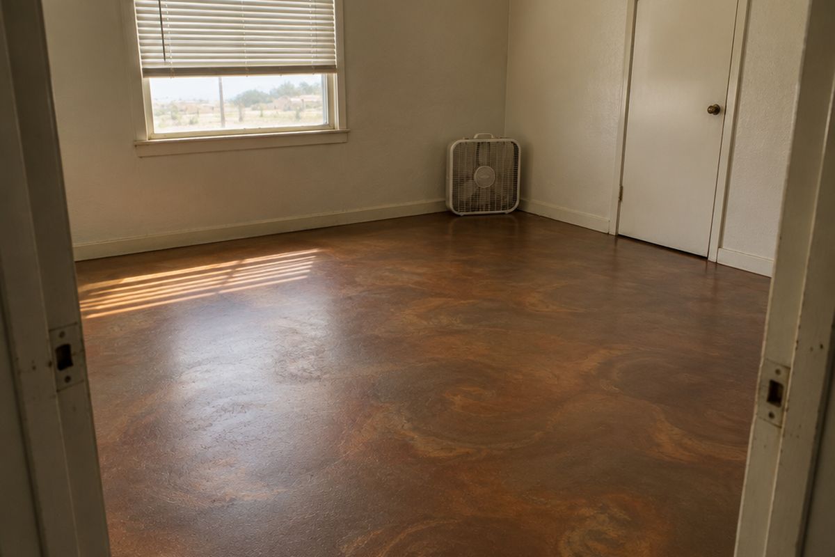 Empty room with a muted bronze and copper-brown metallic epoxy floor, soft stripes of sunlight through half-drawn blinds and a box fan by the wall.