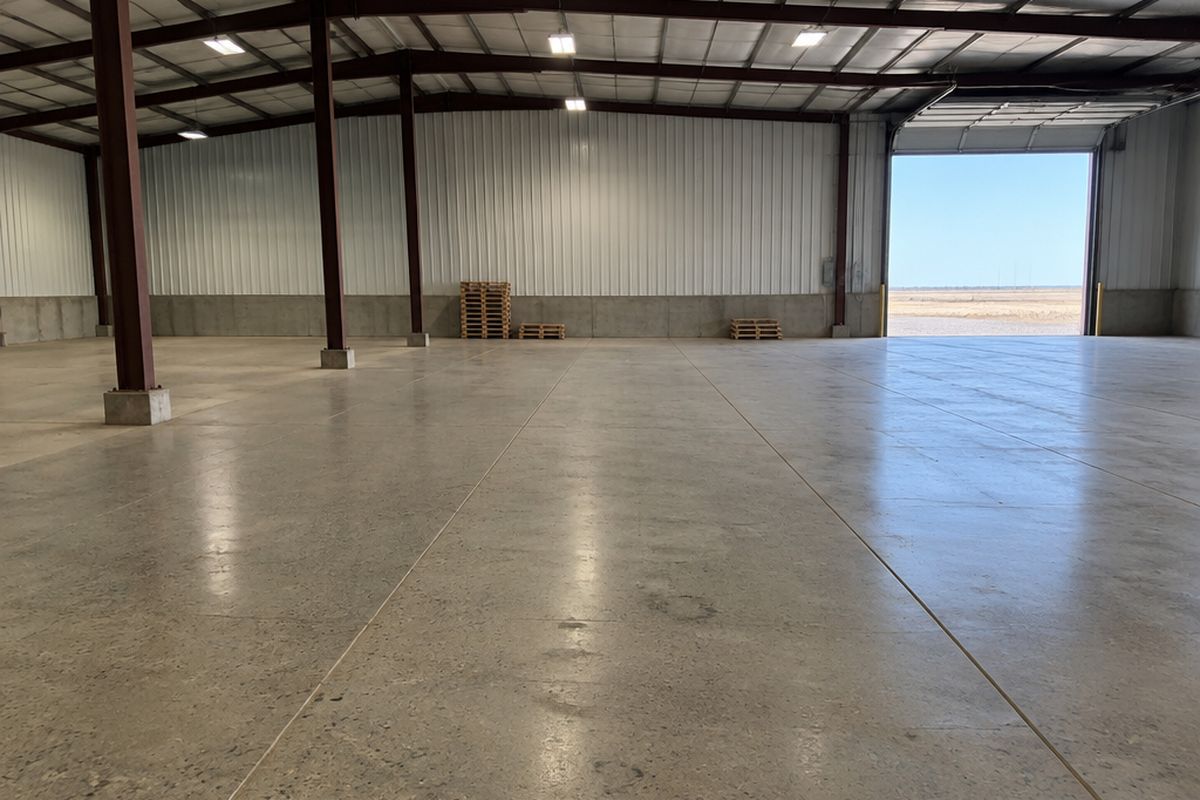 Large, nearly empty warehouse with a polished concrete floor, salt-and-pepper aggregate, long saw-cut joints, steel columns and stacked pallets by the wall.