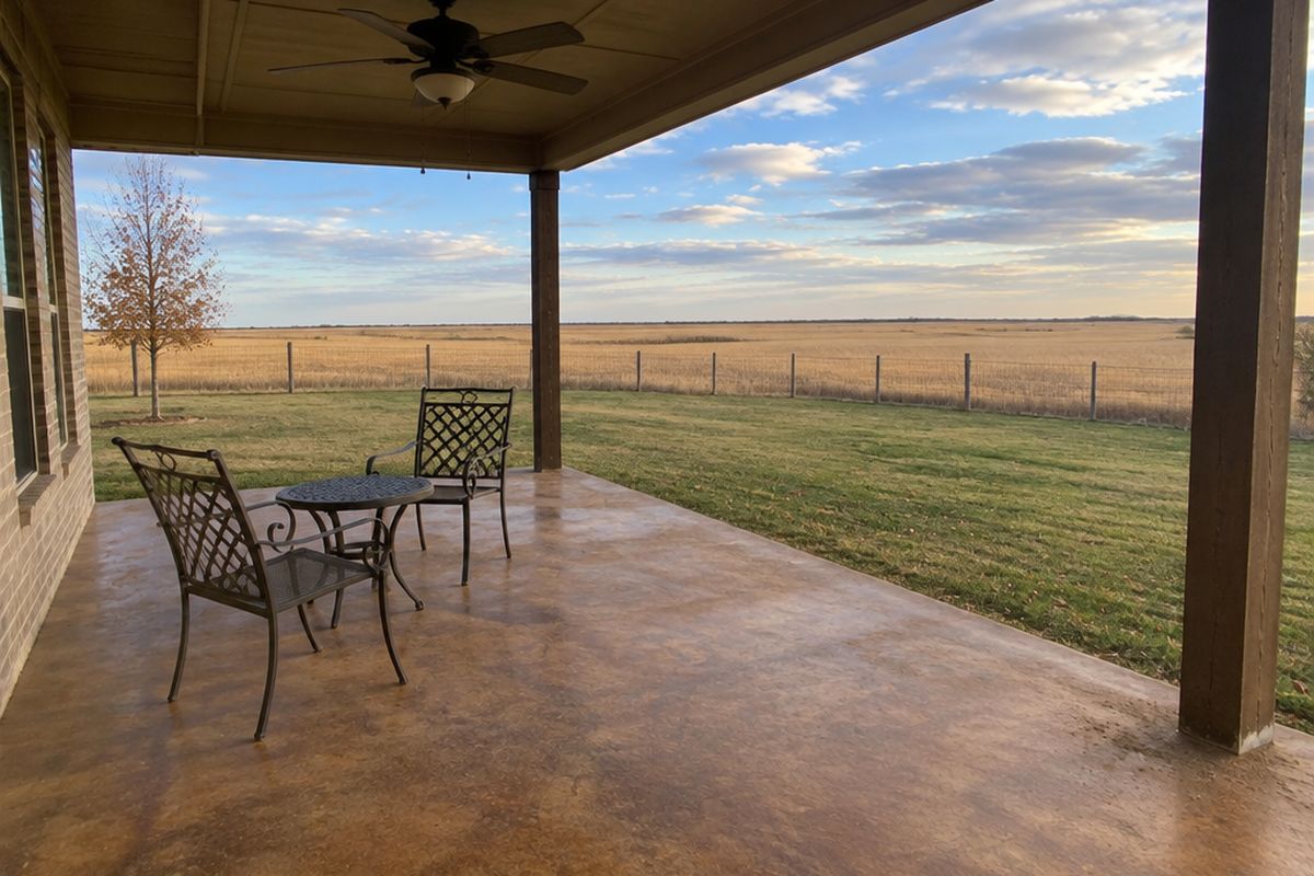 Covered patio with a warm brown and tan stained concrete slab, metal chairs and a small table, looking out over a lawn and a flat wheat field.