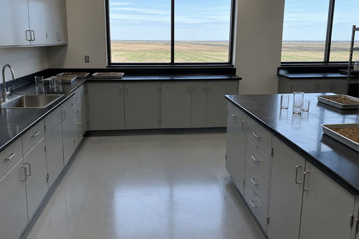Laboratory workroom with a seamless light gray epoxy floor and coved base, black lab benches, a stainless sink and windows onto flat research fields.