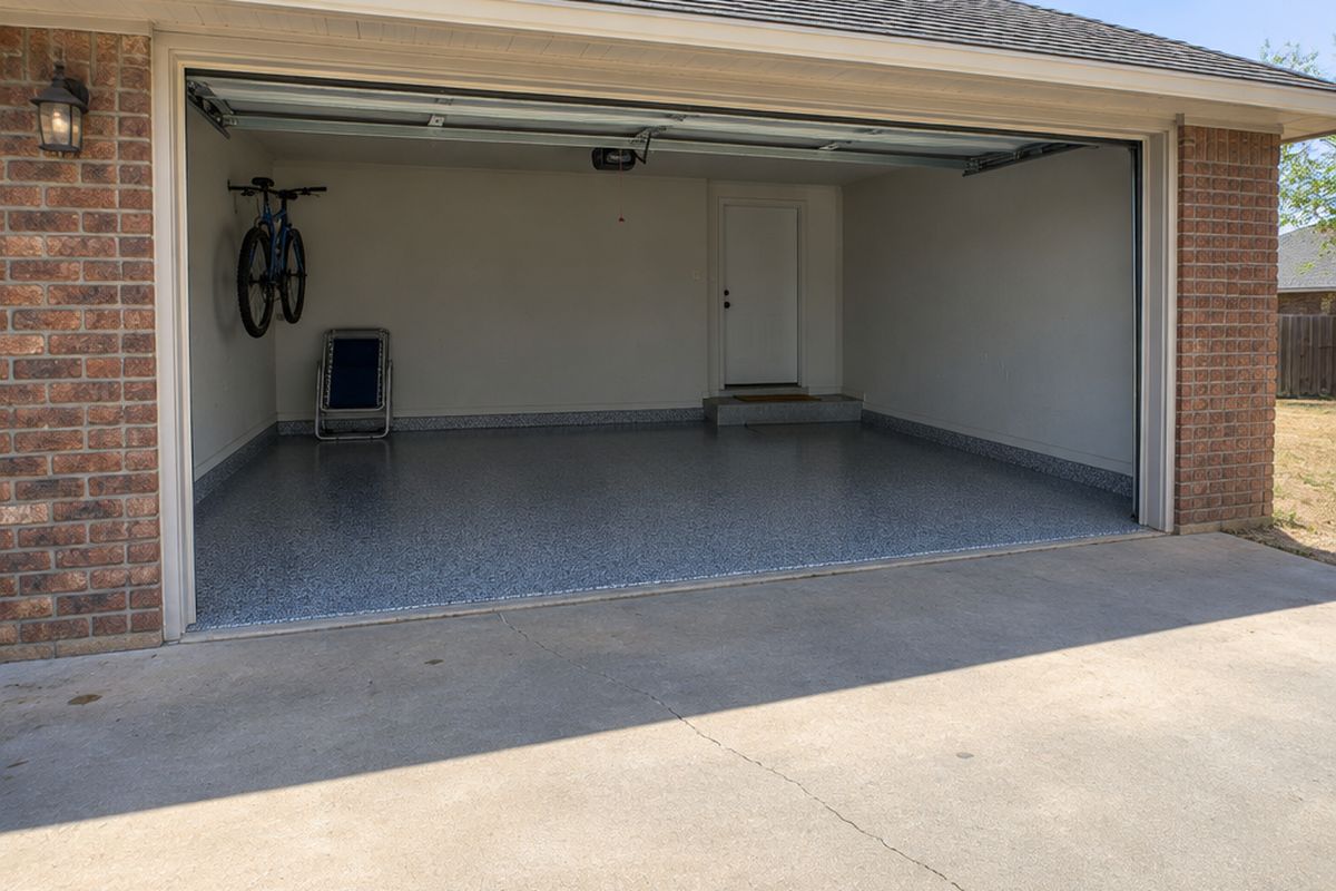 Single-car garage seen from the driveway, its empty blue-gray flake floor in daylight, a bicycle on a wall hook and a step up to the house door.