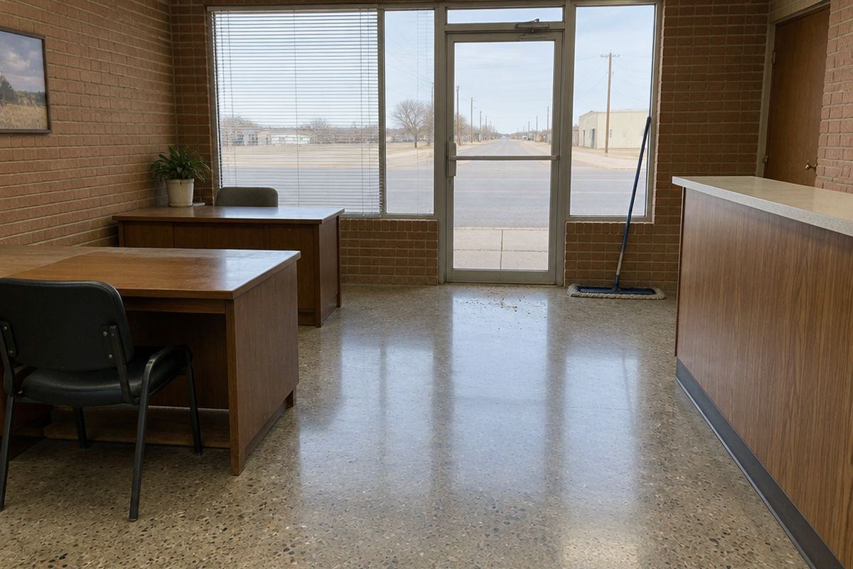 Small office with a medium-sheen polished concrete floor, two wooden desks and a counter, a band of tracked-in grit at the glass door and a dust mop.