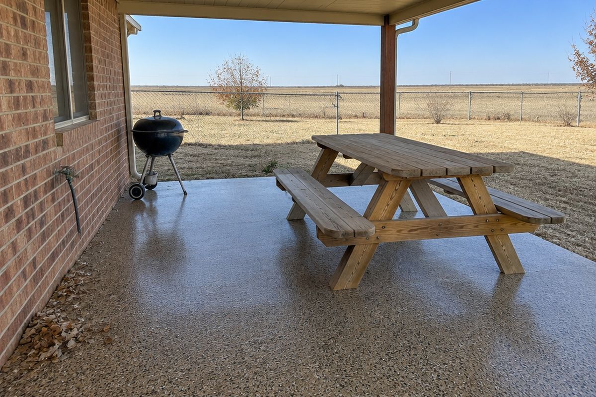 Covered back patio with a tan, gray and black flake coating, a picnic table and grill, a few dry leaves and a flat fenced yard beyond.