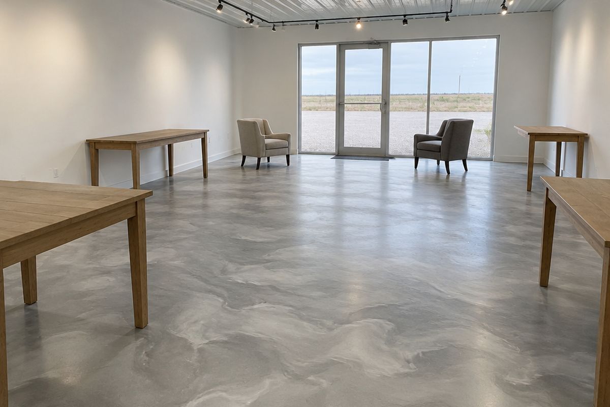 Small showroom with a soft gray and pearl metallic epoxy floor, plain wooden display tables, two chairs and a glass door onto a gravel lot.