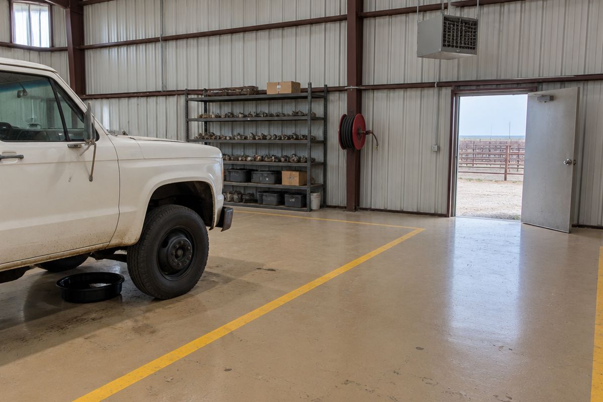 Feedyard maintenance shop with a tan epoxy floor and yellow aisle stripes, an old white truck over a drain pan, a parts shelf and a hanging heater.